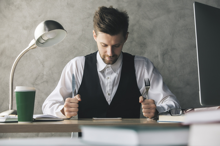 Crazy businessman about to eat cellphone with a knife and fork while sitting at wooden office desk with coffee cup and other items